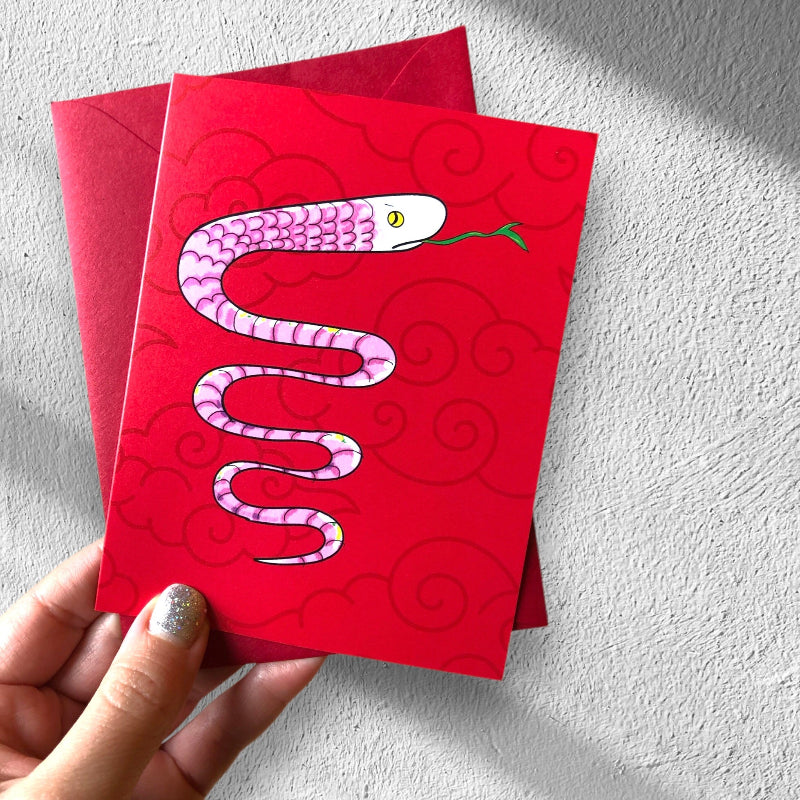 year of the snake card in hand, with matching red envelope, against a white drywall. the card features a pink snake