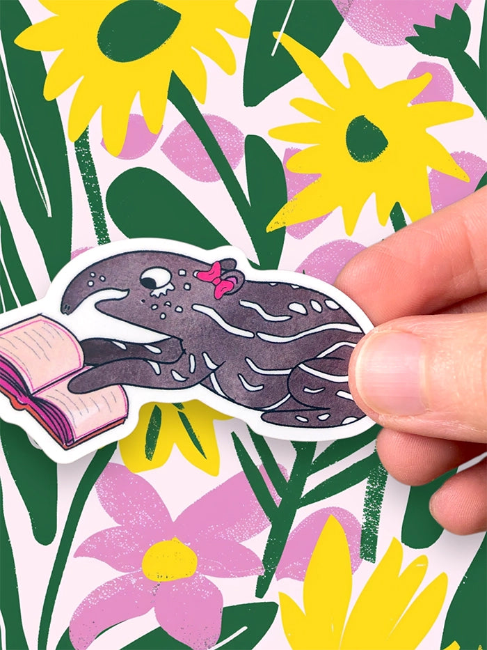 Hand holding a sticker of a gray baby tapir with a pink bow against a floral background