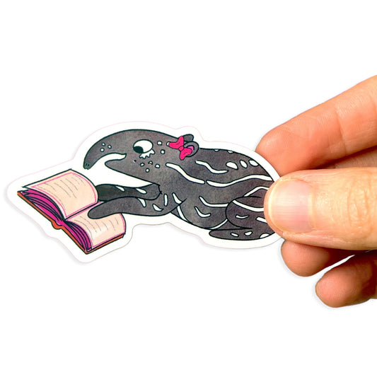 Hand holding a sticker of a baby tapir with a pink bow reading a book on a white background