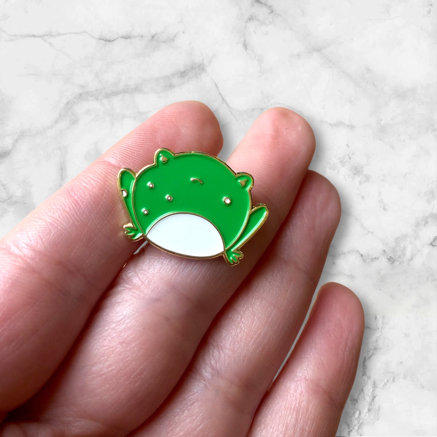 angry but cute frog pin in hand, against a marble background
