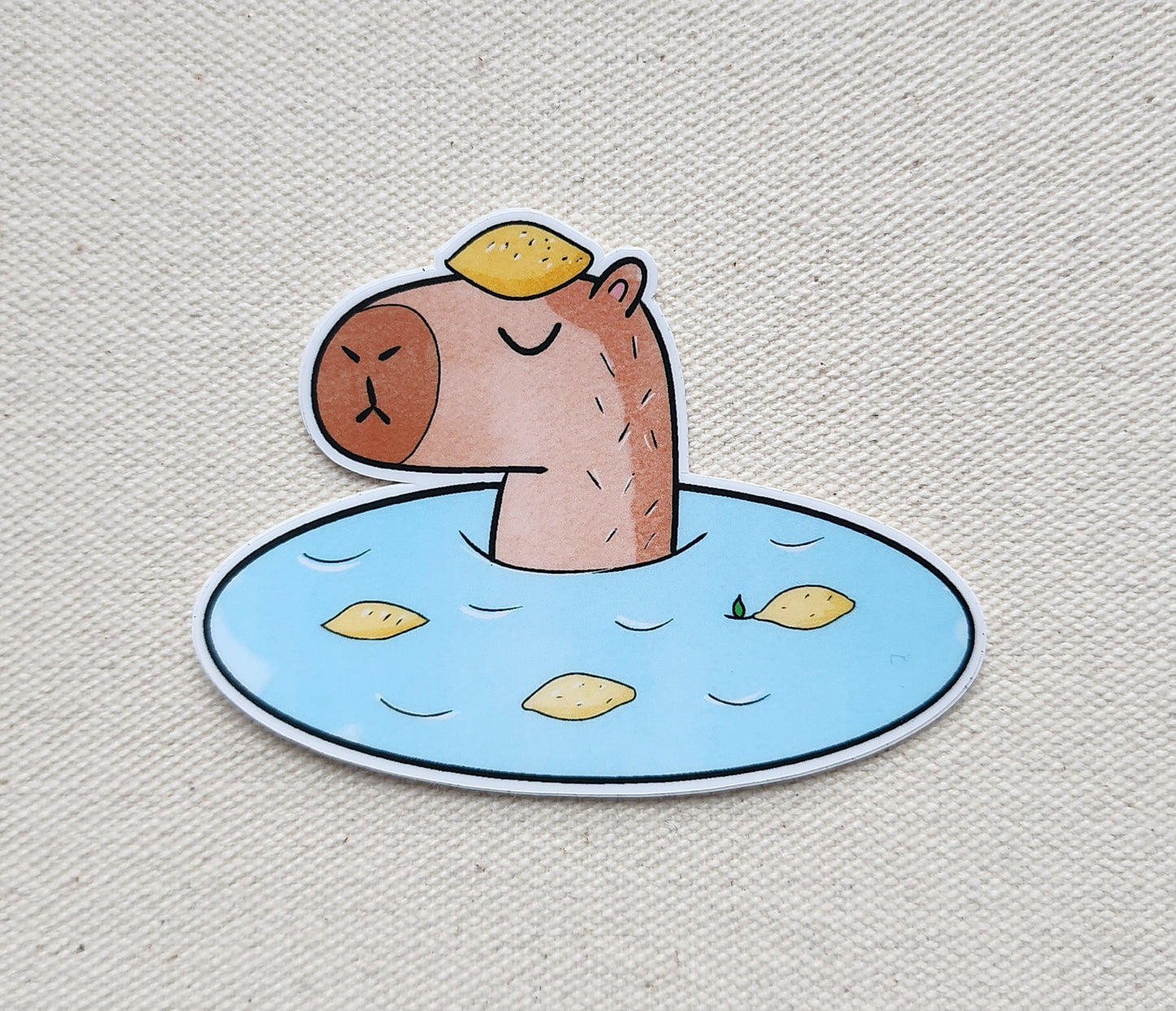 Sticker of a capybara in an onsen with a lemon on its head, against a beige fabric background
