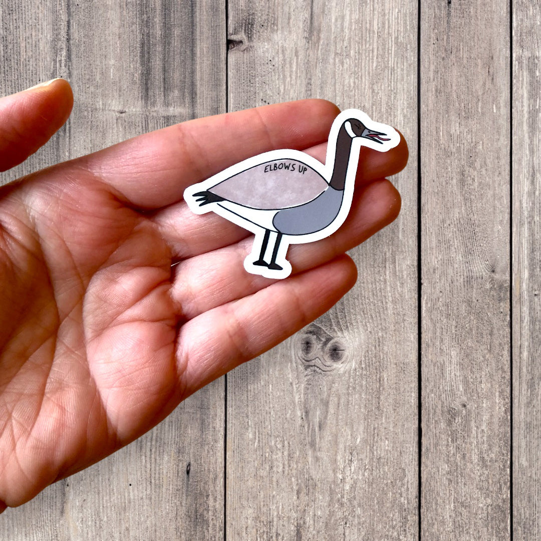 elbows up canada goose sticker in hand, against a wooden background. perfect gift for bird lovers, or wanting to show your canadian pride