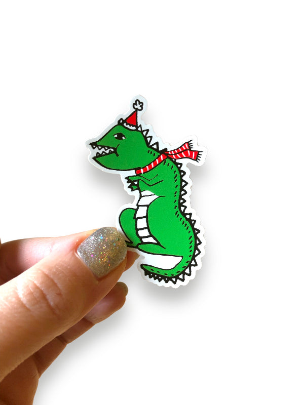 dino vinyl sticker decked in red santa hat and red scarf with white stripes, in hand against red background