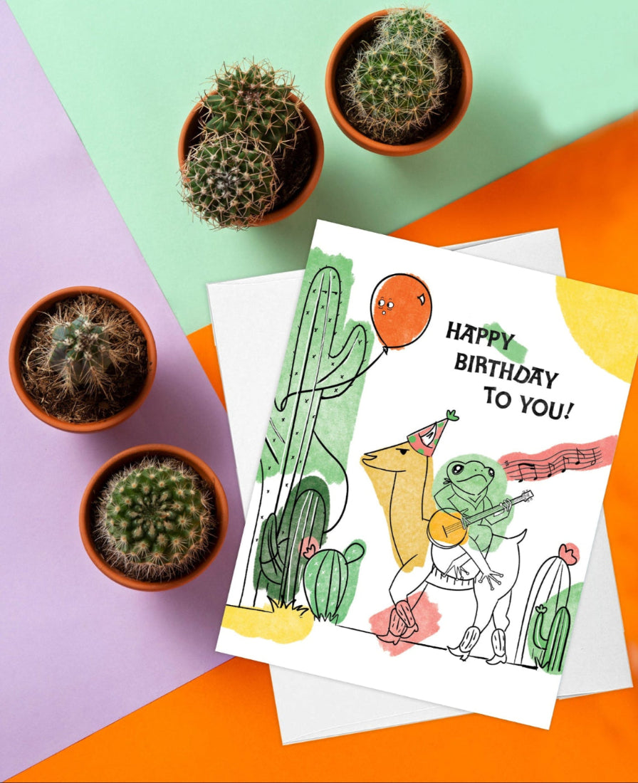 a cute and fun birthday card featuring a frog riding a llama, playing the banjo and singing happy birthday. perfect for any frog or llama lover (or both). the card is placed against a colourful background with cacti plants
