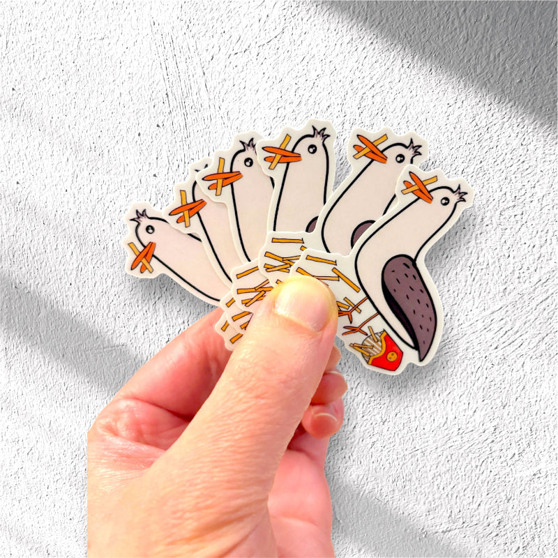 Hand holding a bunch of stickers of Gus the Gull eating fries against a light gray background