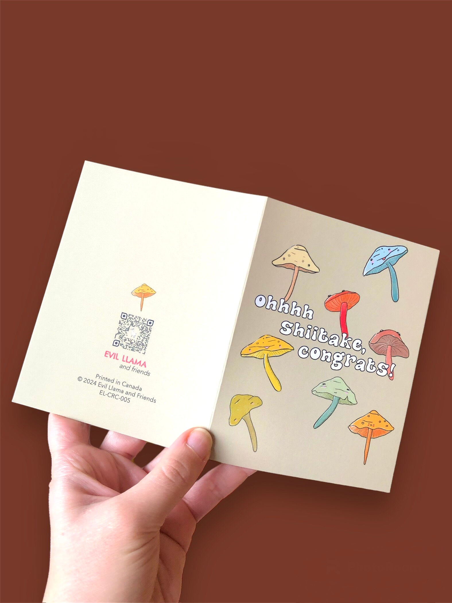 "oh shiitake congrats" card in hand, showing front and back of the card. the front has a bunch of shiitake mushrooms in earth tones, with the text reading "ohhh shiitake, conrats!", the back shows our brand with logo and QR code. the card is held against a brown background