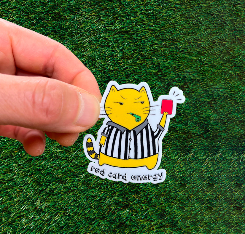 Sticker of a grumpy cat in a referee outfit holding a red card on green grass.
