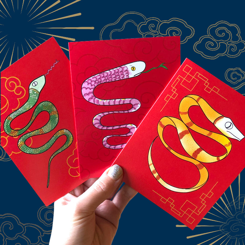 3 lunar new year cards in hand against a blue background. each card features a different snake, one green, one pink, one yellow