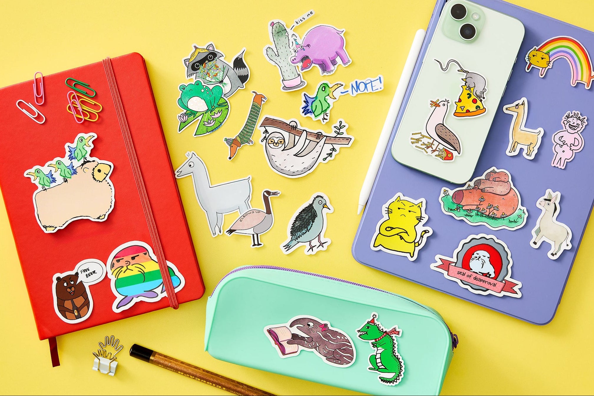 A styled flat-lay of colourful animal-themed stickers on a yellow background, shown applied to a red notebook, a green phone case, a purple iPad, and a mint green pencil case, with pencils and paper props around them.
