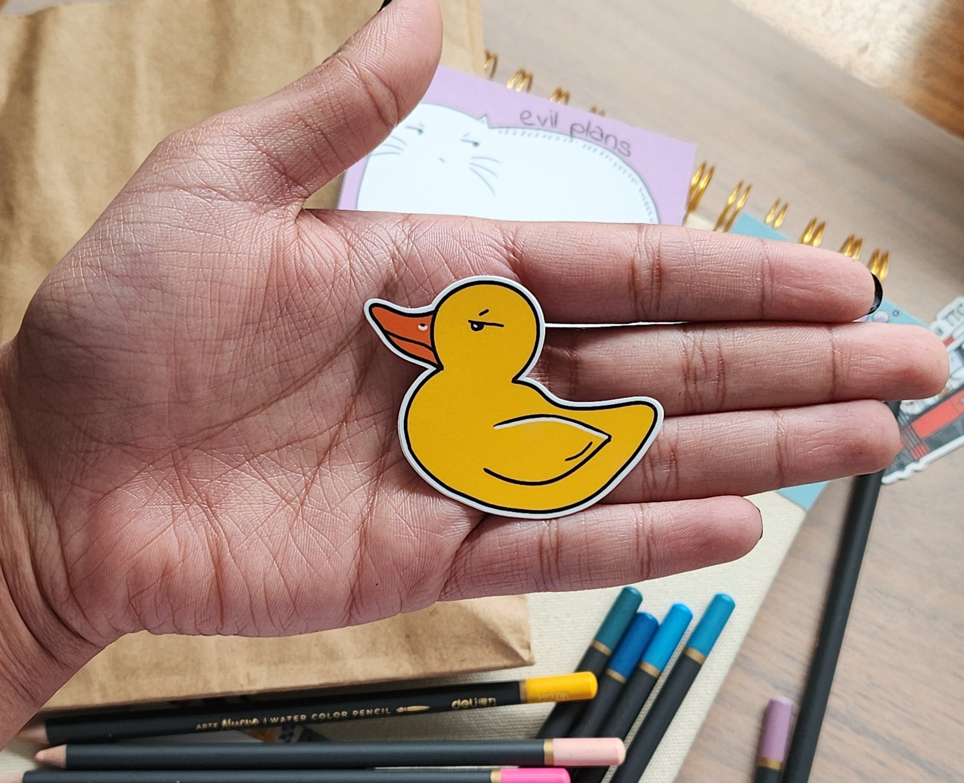 Hand holding a yellow rubber duck sticker with stationery items in the background