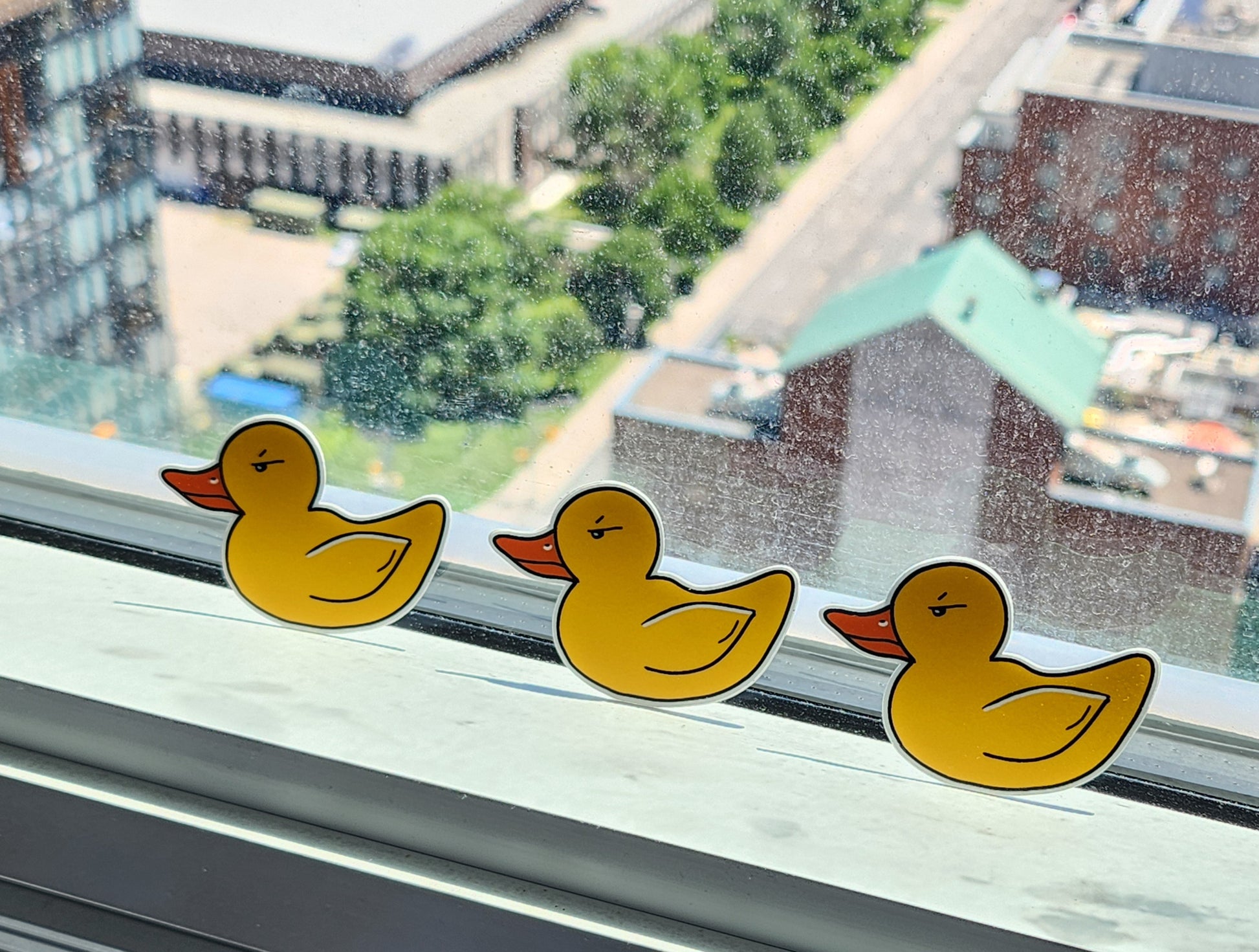 Three yellow rubber duck stickers on a window with a cityscape background