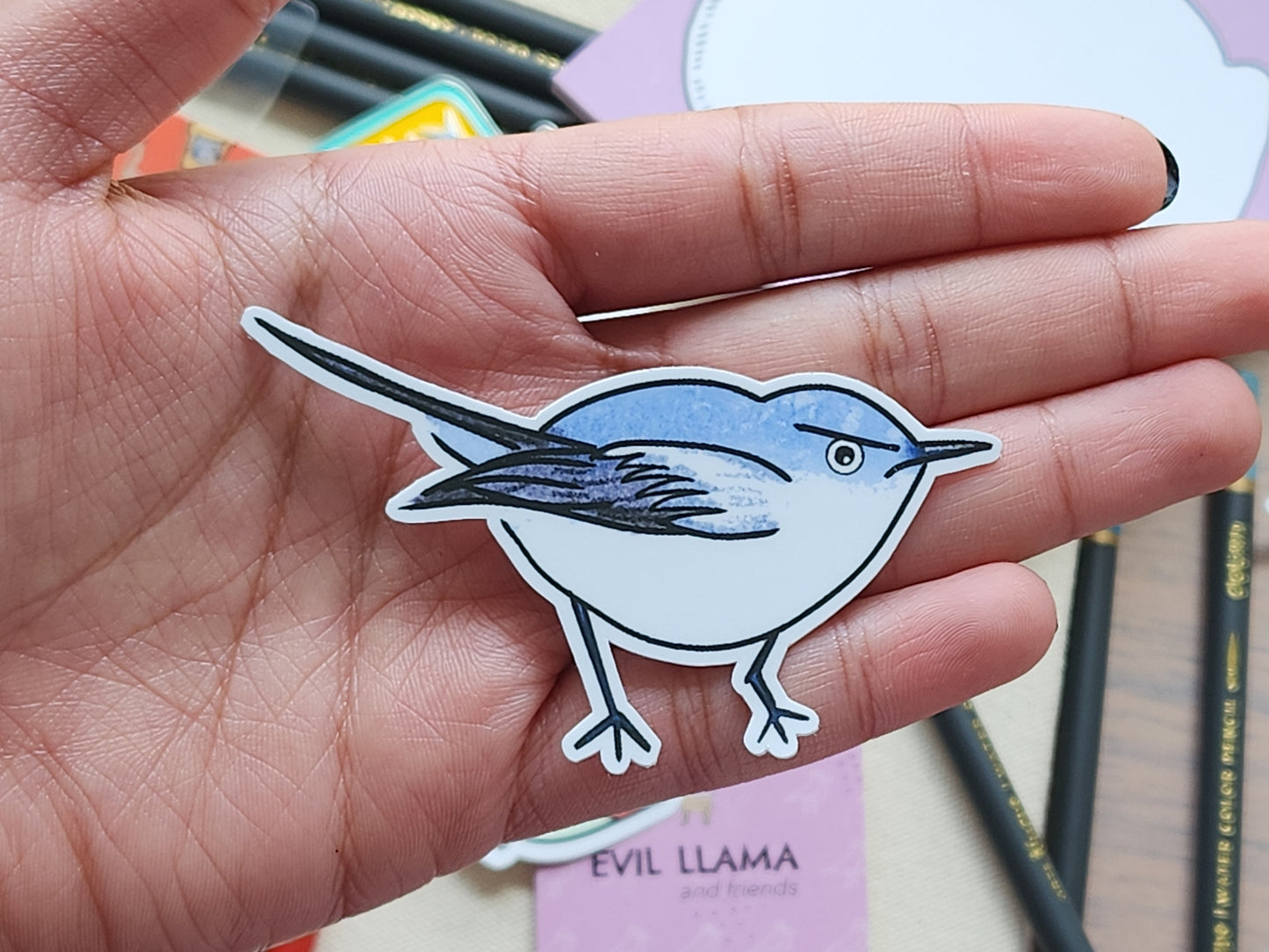 Hand holding a gnatcatcher bird sticker with stationery items in the background