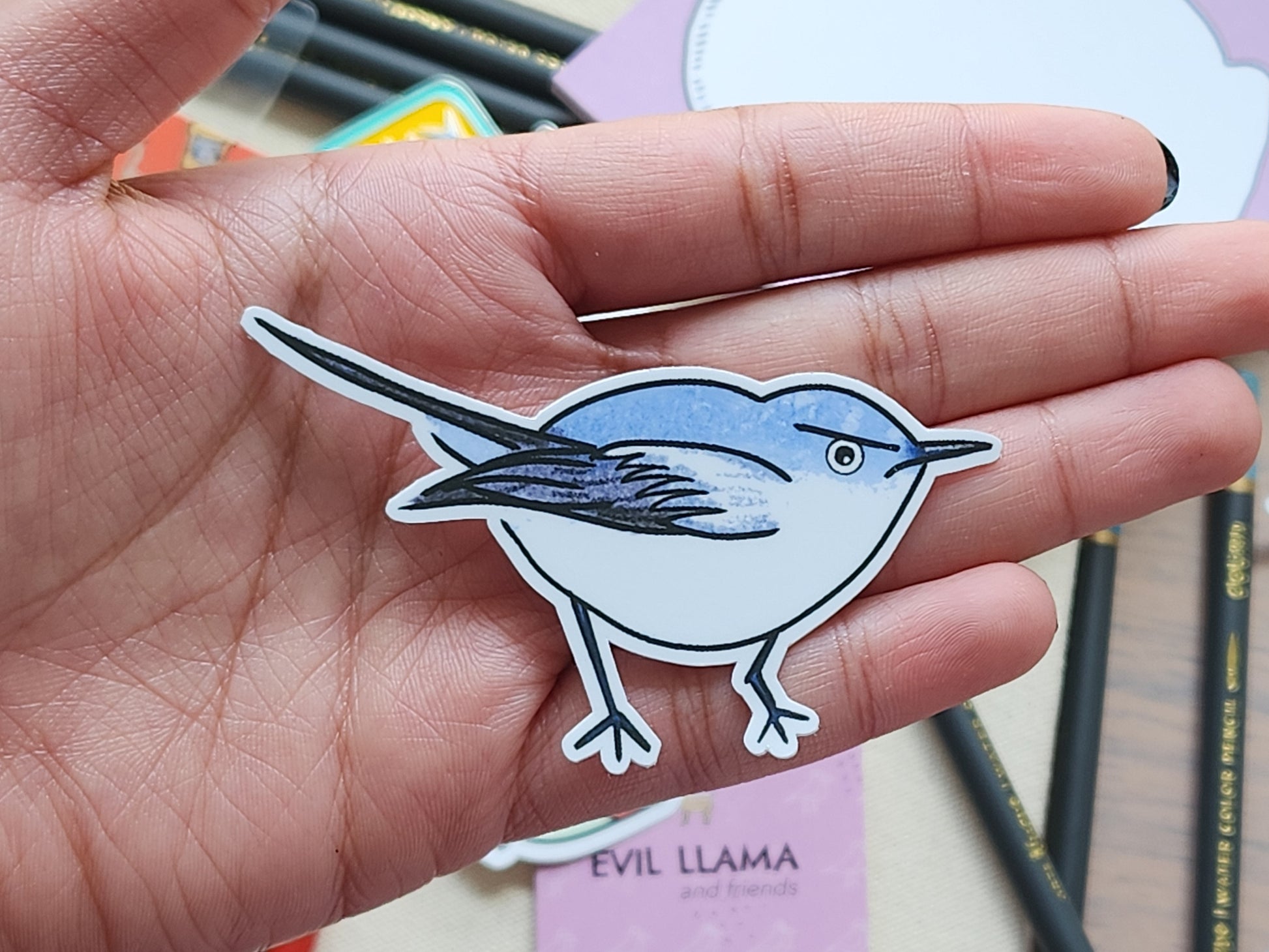 Hand holding a gnatcatcher bird sticker with stationery items in the background