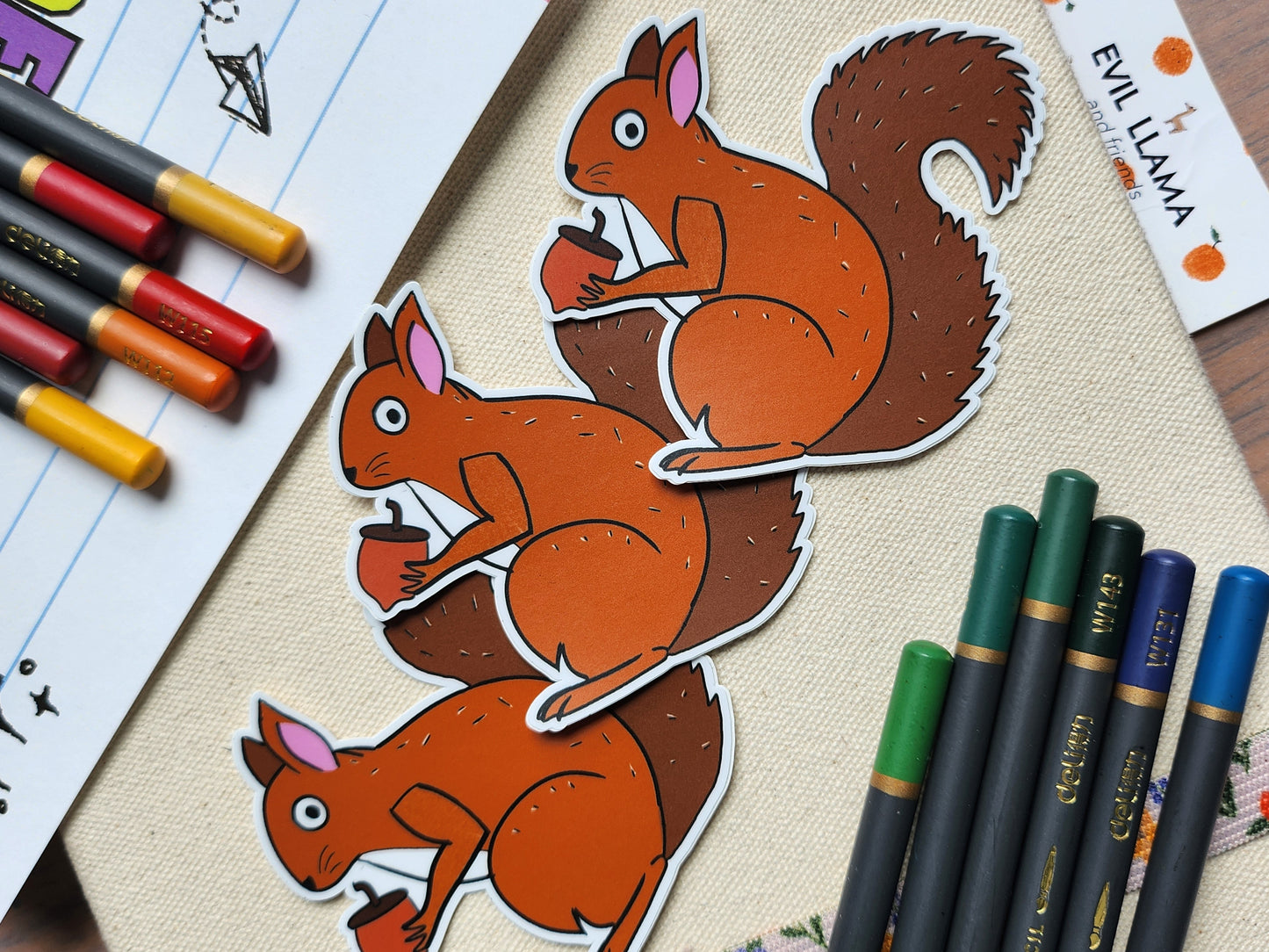 3 squirrel stickers styled next to some coloured pencils, on a beige textured background