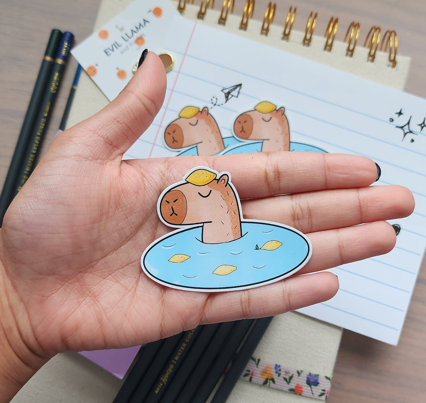 Hand holding a cartoon capybara in onsen sticker with a background filled with stationery items