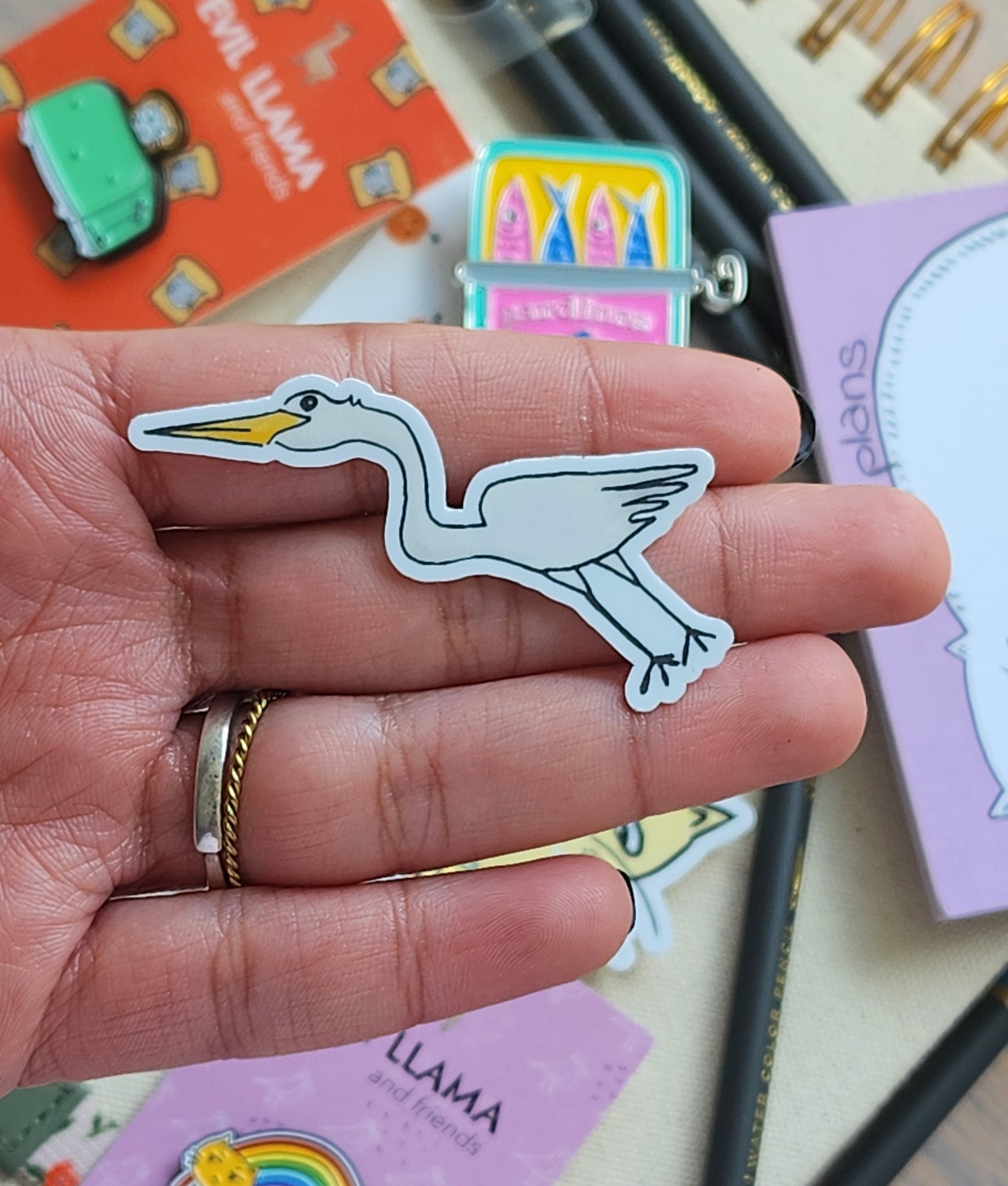 Hand holding a sticker of an egret, surrounded by enamel pins and stationery items.