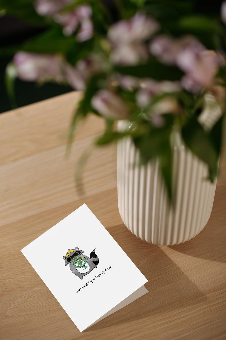 sorry everything is trash raccoon sympathy card, placed on table next to flowers. the perfect card to let someone going through a tough time that you are there for them in a time of need