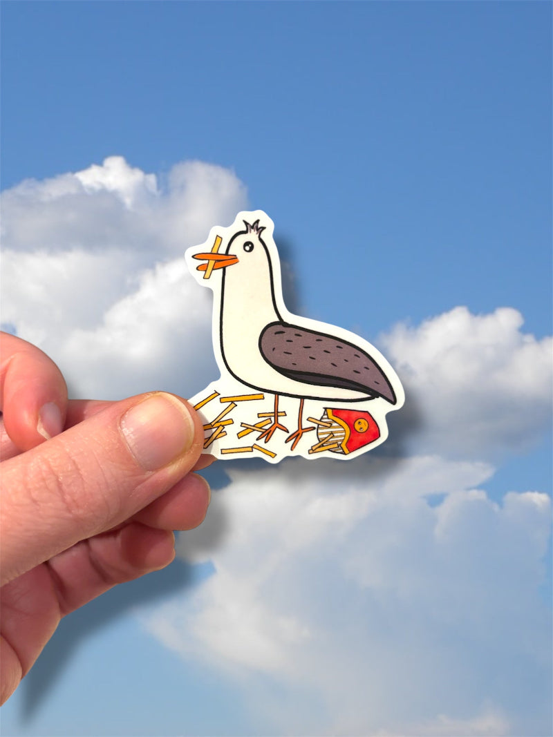 Hand holding a sticker of a cartoon seagull eating fries against a blue sky with clouds