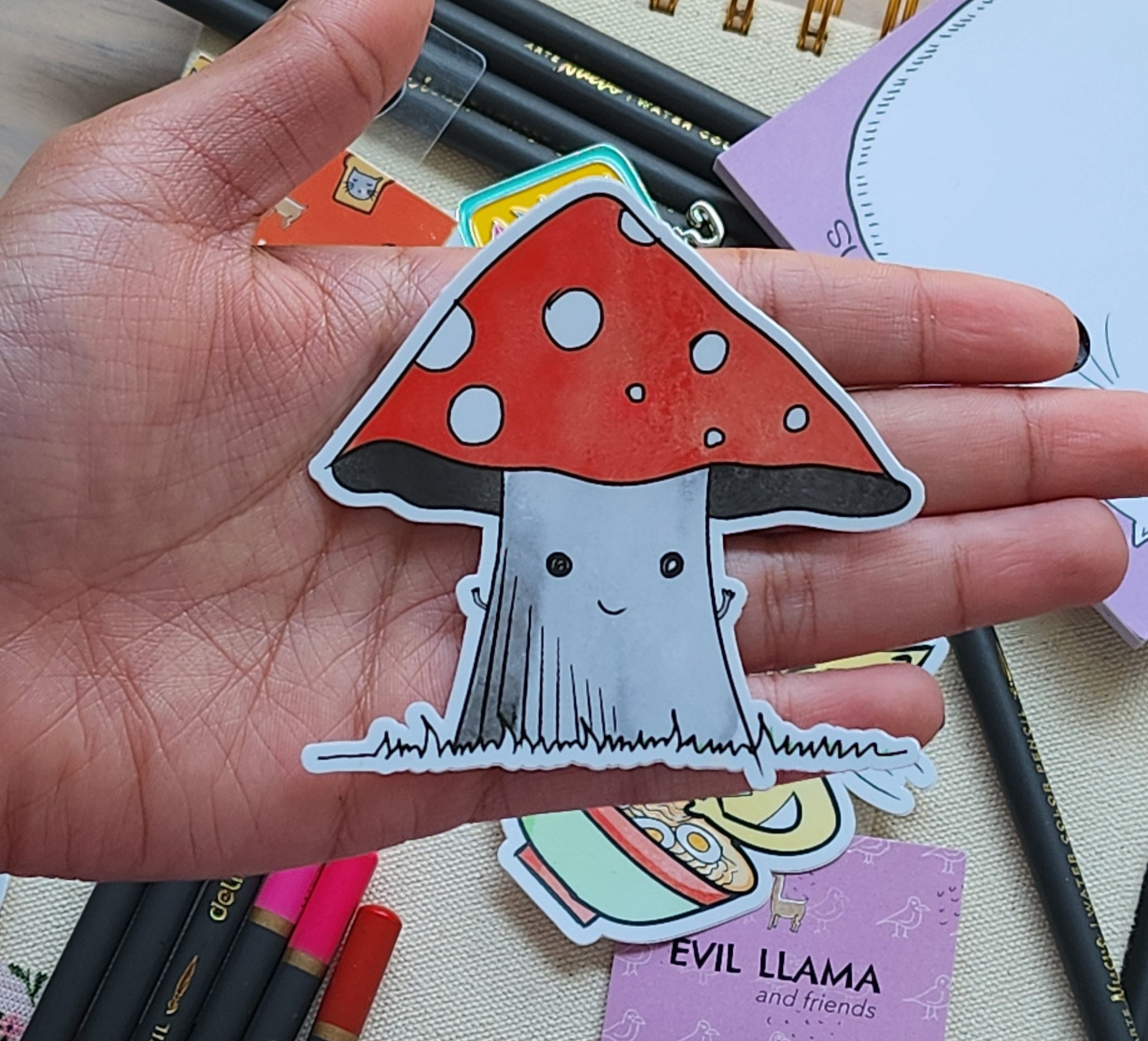 Hand holding a mushroom-shaped sticker with a red cap and white spots, surrounded by stationery items.