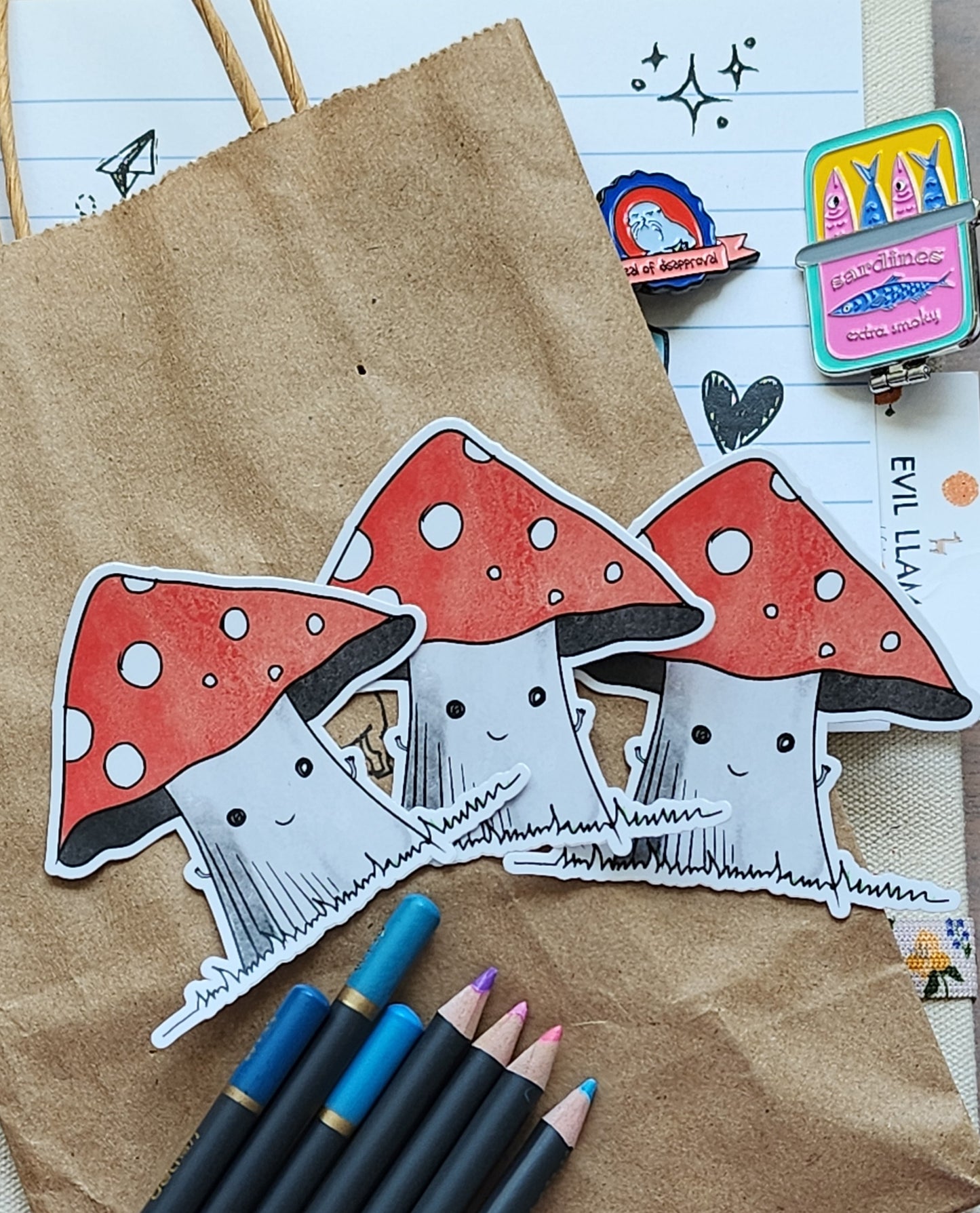 3 happy mushroom stickers on a craft paper bag, surrounded by colouring pencils and some enamel pins