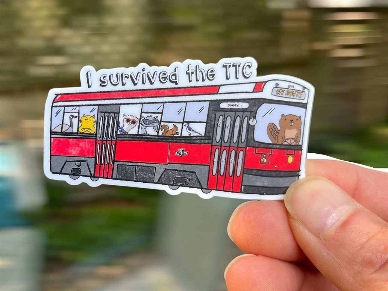 sticker showing a toronto streetcar (TTC) packed with animal passengers, in hand, against a blurry background indicating motion. the text on top reads "I survived the TTC"