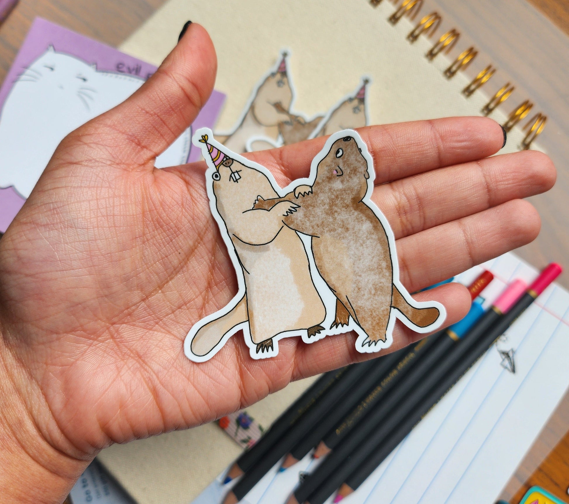 hand holding a sticker featuring two marmots, against a background of various stationery items