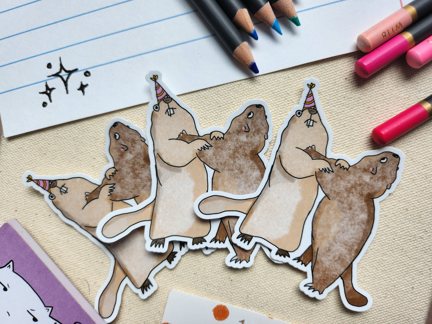 3 marmot stickers styled next to some colouring pencils, on a textured beige background