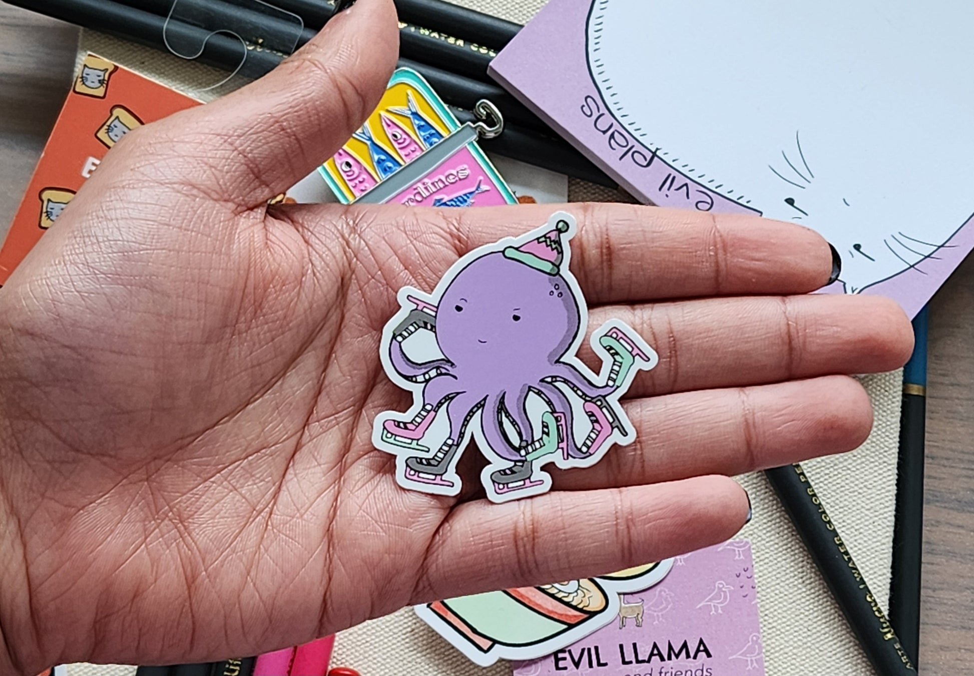 Hand holding a purple octopus sticker wearing skates, against a colourful background.