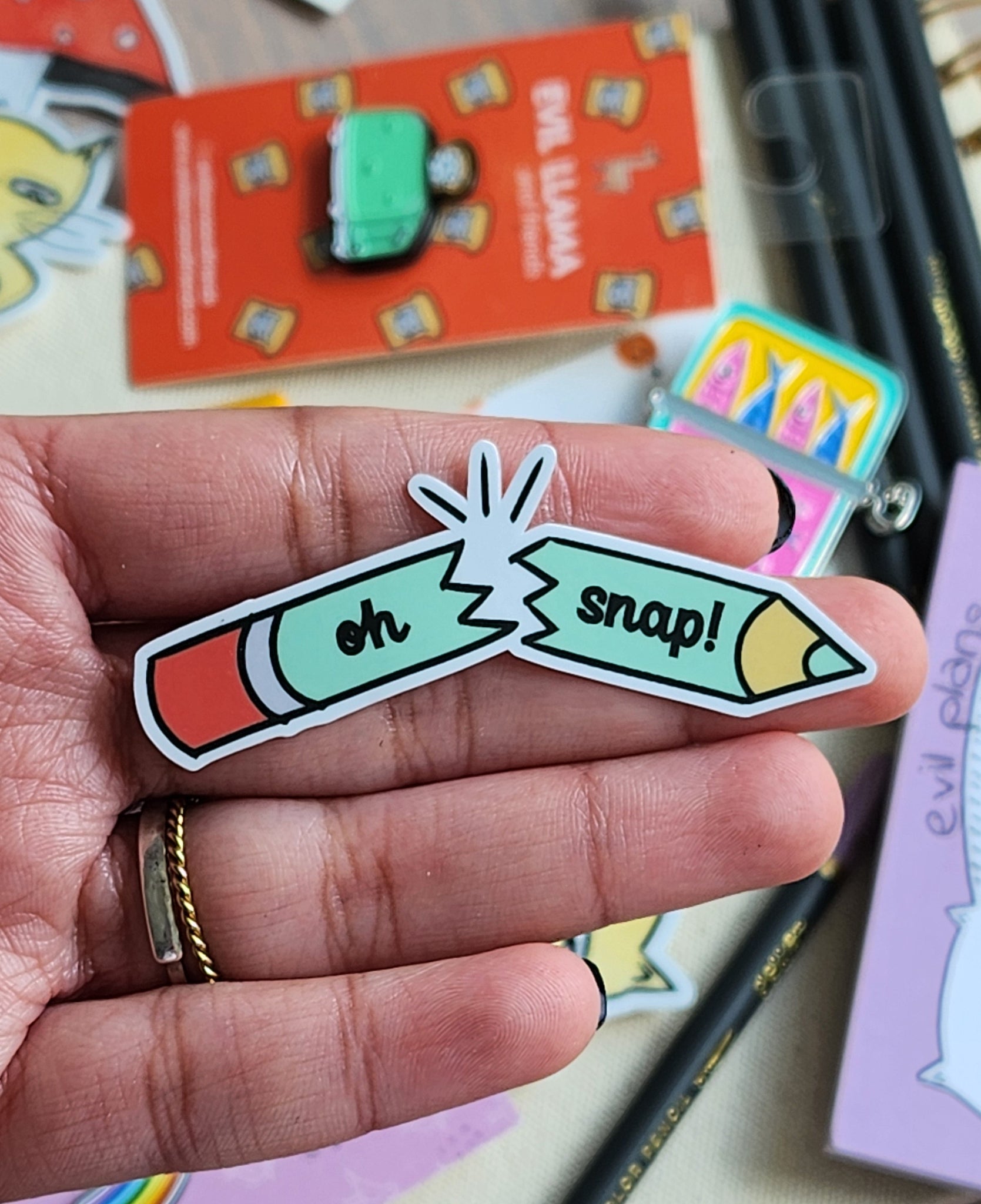 Colorful 'oh snap!' pencil-shaped sticker held in a hand with a blurred background of stationery items.
