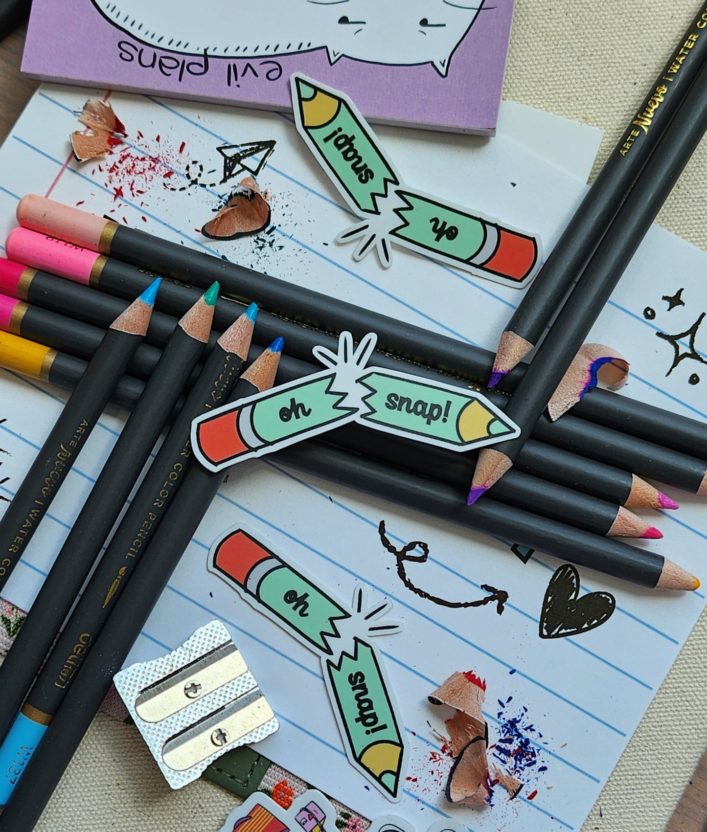 "oh snap" pencil stickers on coloured pencils, styled next to a pencil sharpener, lined paper with doodles, and a small notepad.
