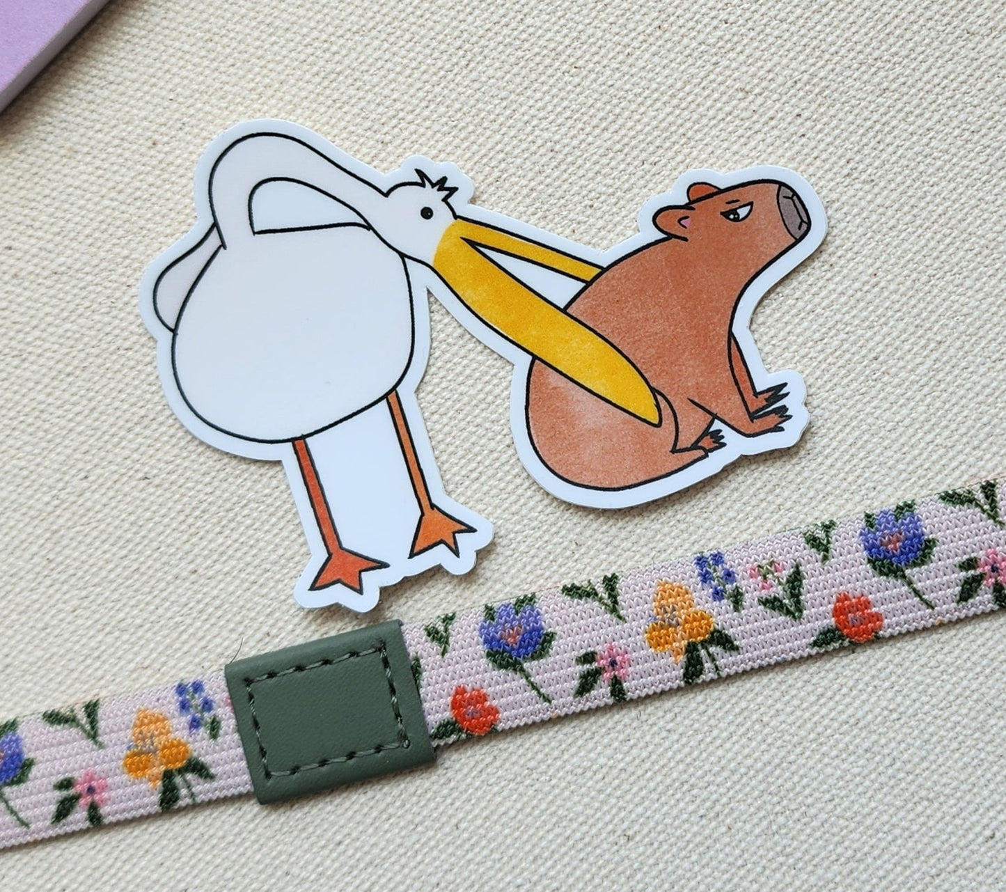 pelican eats capybara sticker on a textured background, with a floral band underneath