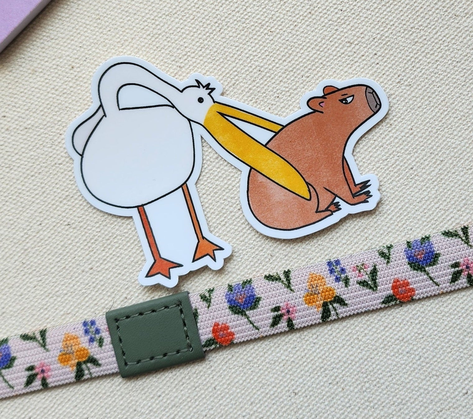 pelican eats capybara sticker on a textured background, with a floral band underneath