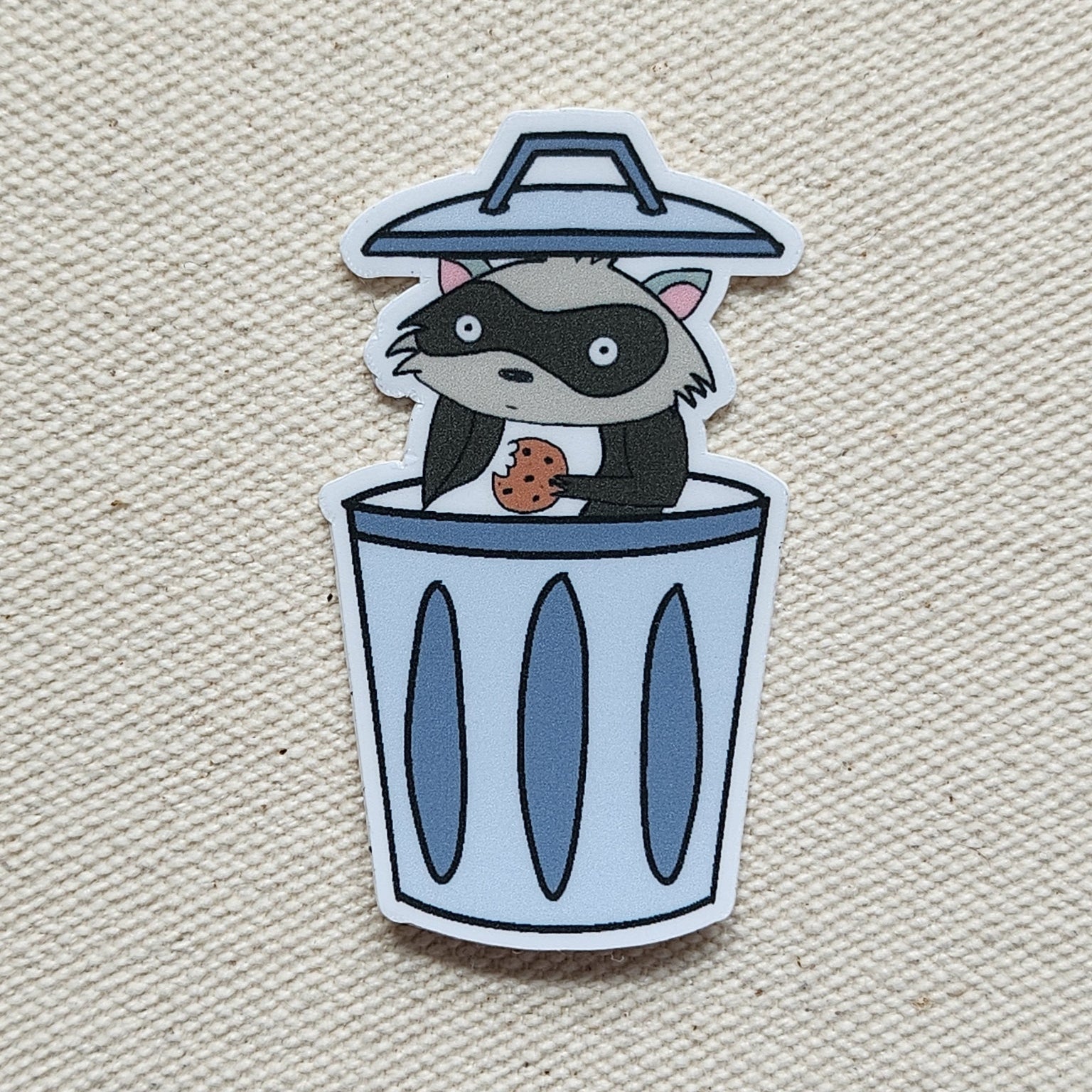 raccoon sticker in garbage can against a beige, textured background