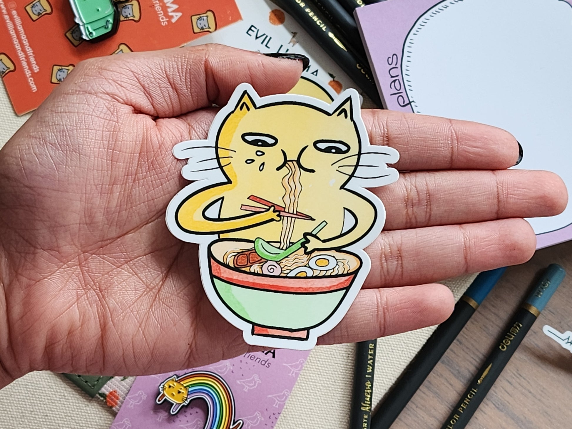 hand holding a sticker showing a cat slurping down a bowl of ramen. the background is filled with various stationery items