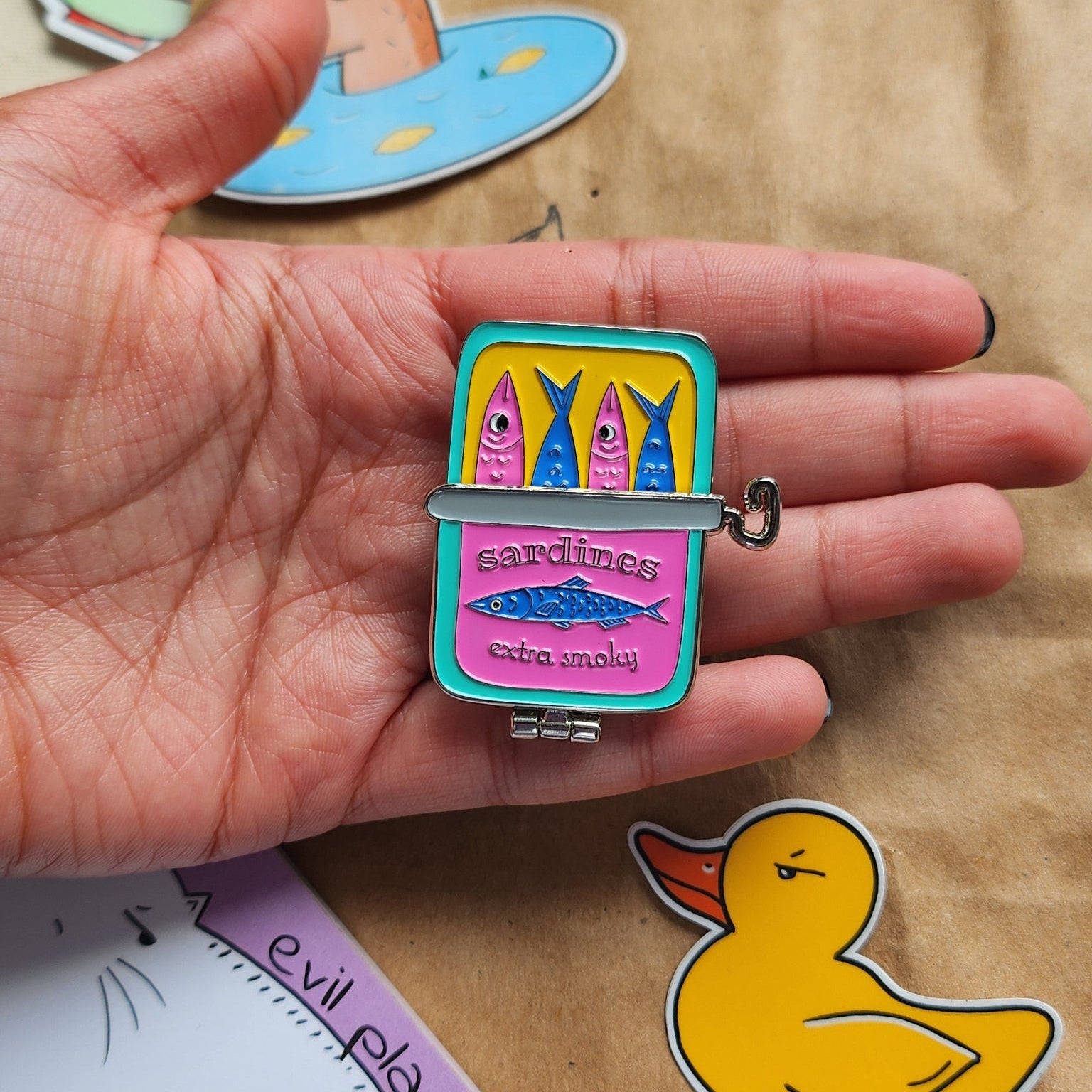hand holding a pin featuring a canned sardine with a hinge