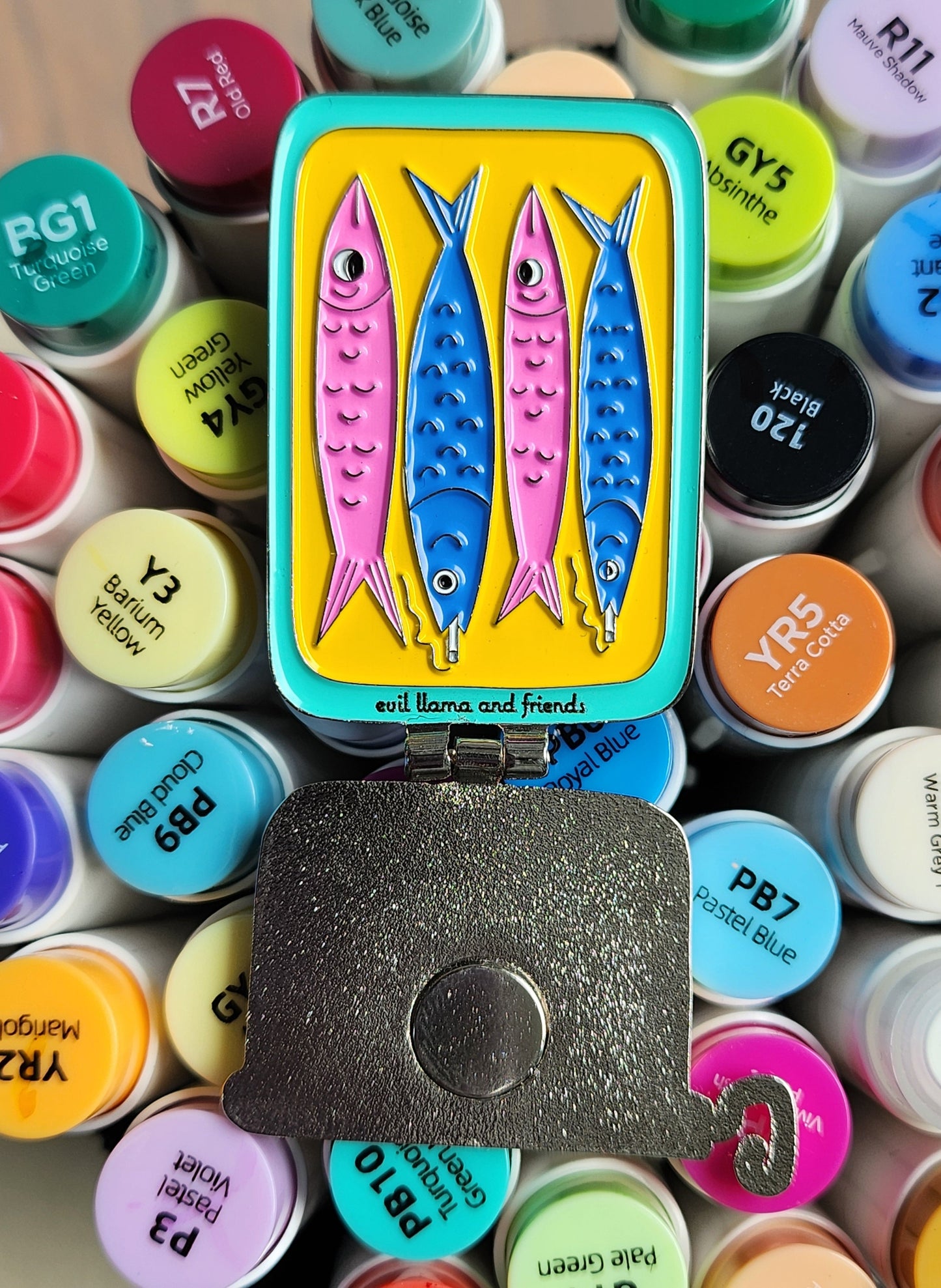 sardine can pin in open position, showing the inside with two smoking fish, on top of colourful markers
