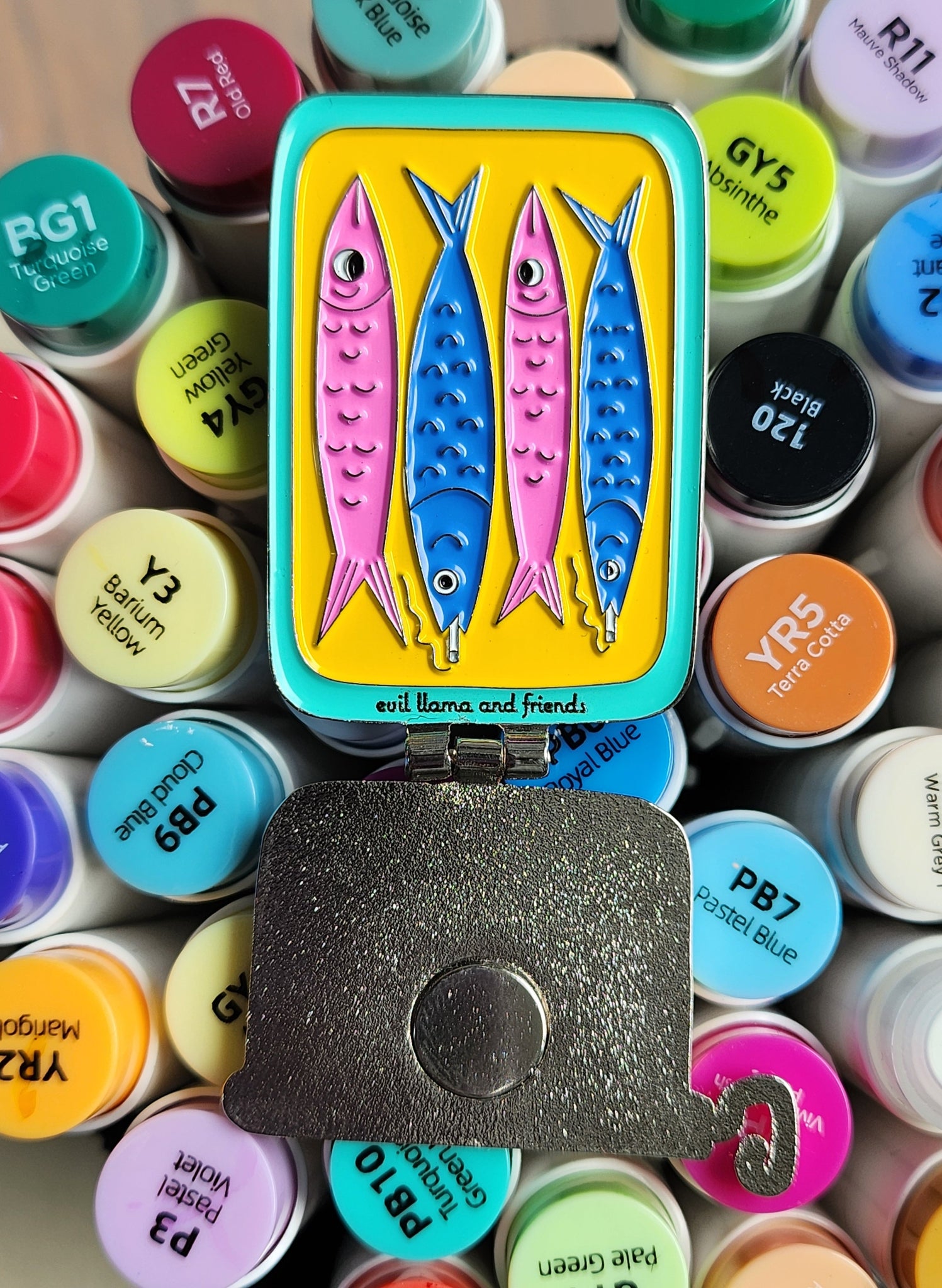 sardine can pin in open position, showing the inside with two smoking fish, on top of colourful markers