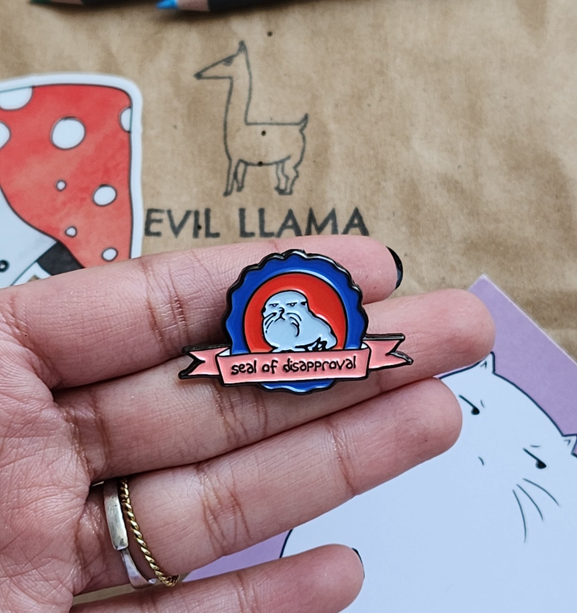 hand holding a pin featuring a grumpy seal, with the text reading "seal of disapproval". there is a craft paper bag and some stationery items in the background