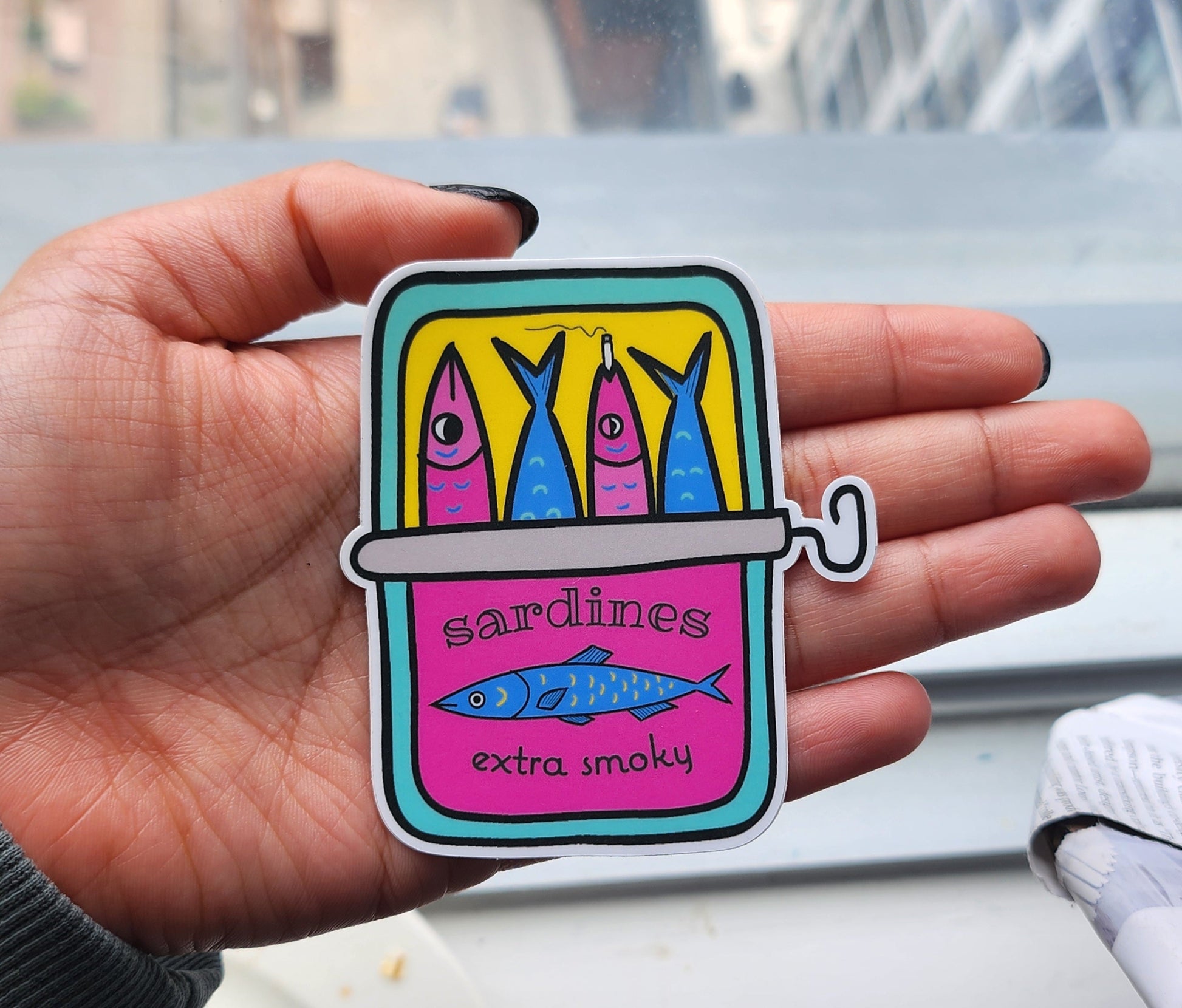 hand holding a sticker featuring a tin of sardines. the text reads "sardines extra smoky", and one of the sardines is having a smoke!