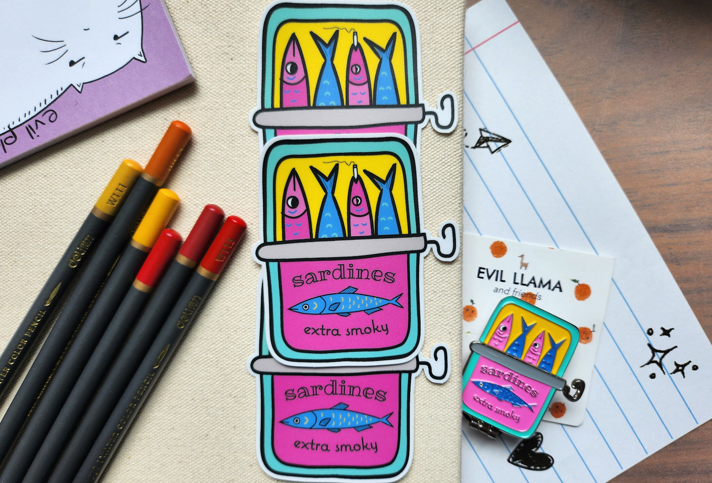 3 smoky sardine stickers styled next to some colouring pencils, notepad, and an enamel pin