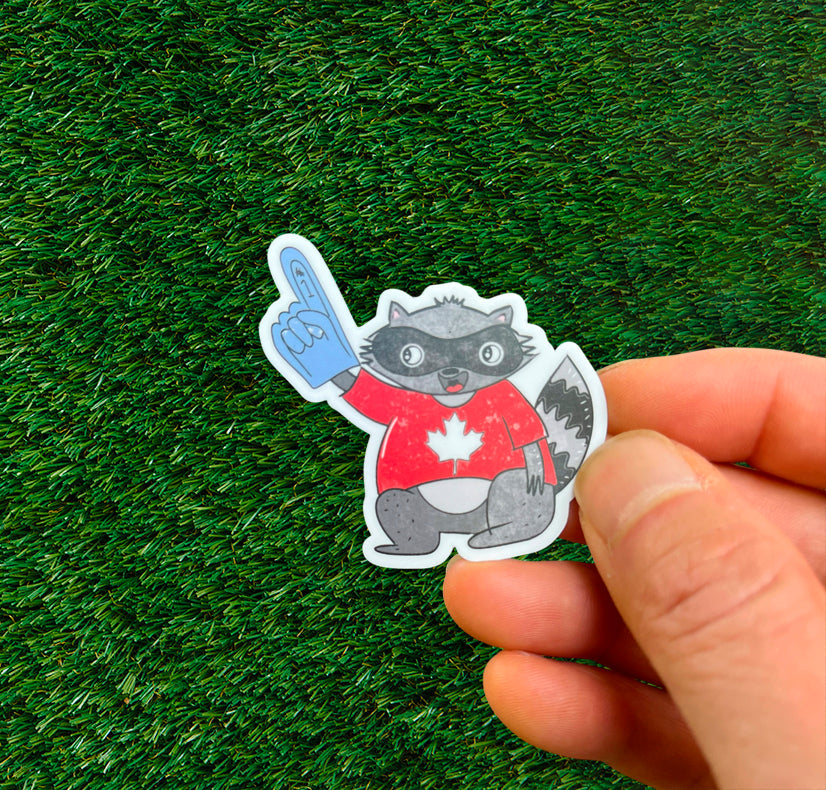 Sticker of a raccoon wearing a red Team Canada jersey with a maple leaf, and blue foam finger, on green grass